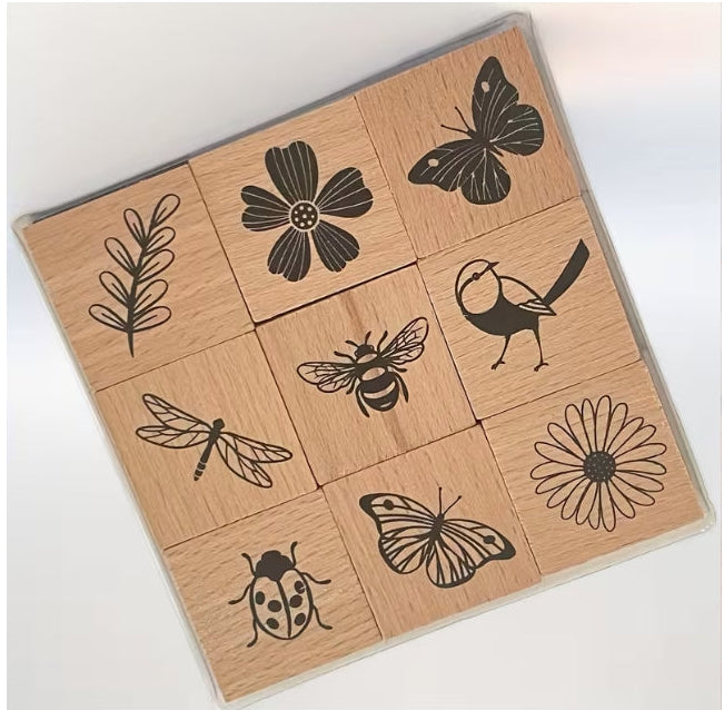 A 3x3 set of wooden stamp blocks in a clear plastic case, featuring black engraved illustrations of a daisy, butterfly, ladybug, dragonfly, bird, bee, leaf, and flower.